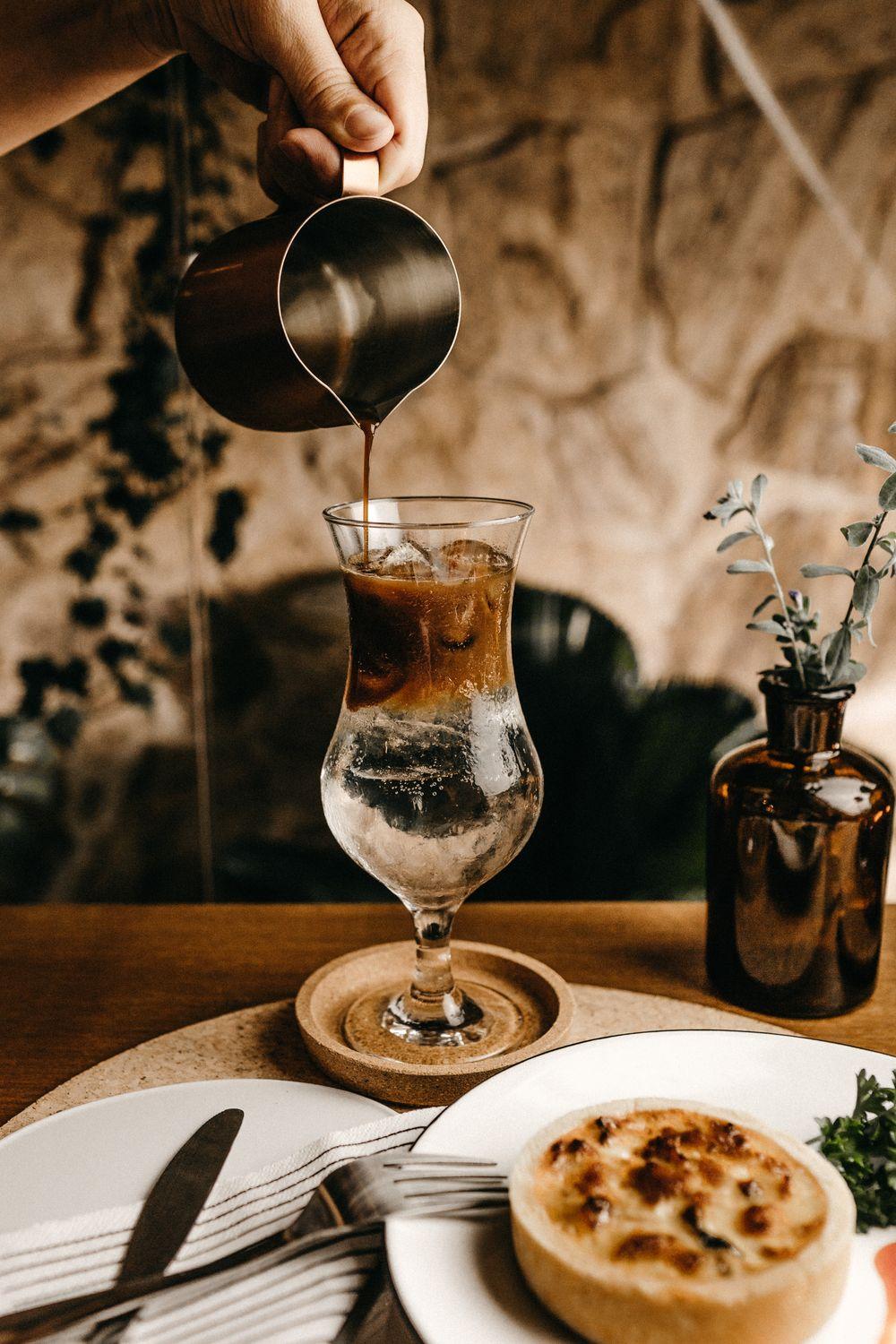 Coffee being poured over ice into a glass in a warm, sunlit cafe